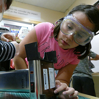 Young student wearing goggles and cutting paper with a paper cutter.