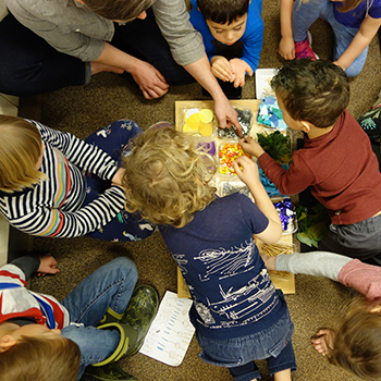 Students sitting in a circle, hovered over various objects sorted into small boxes.