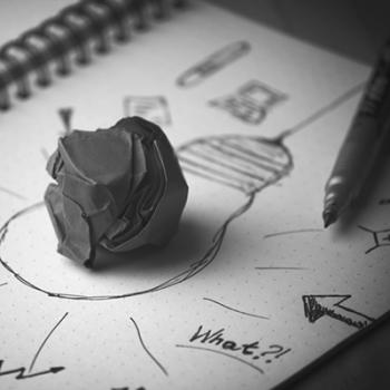 Black and white image of a crumpled paper ball on a notebook with a sketch of a lightbulb.