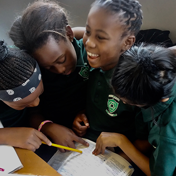 Four young students huddled together around a school book, engaged in a collaborative activity.